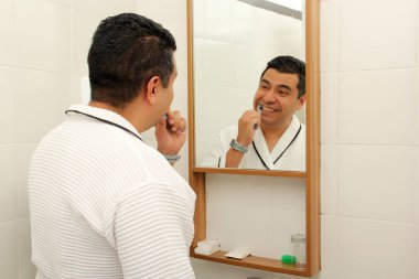 Esmer tenli 40 yaşındaki Latin adam diş dökülmesini ve çürükleri önlemek için banyo aynasının önünde dişlerini fırçalıyor.