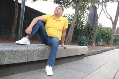 40 yaşındaki esmer tenli Latin adam, rahat ve sakin bir şekilde bankta oturmaktan zevk alıyor. Yeni beyaz tenis ayakkabılarını moda modasında gösteriyor.