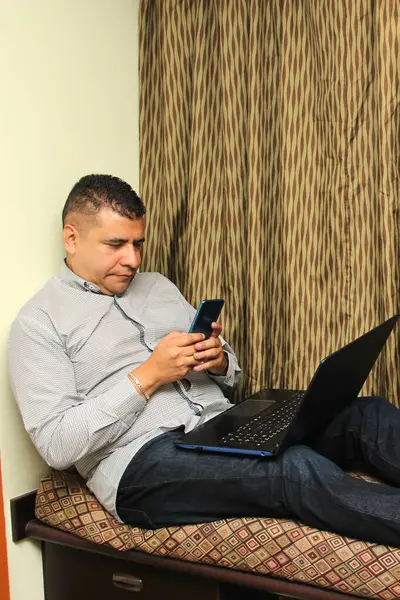 40-year-old dark-skinned Latino man uses his laptop and cell phone to work, make video calls, send messages, make appointments, buy and pay online