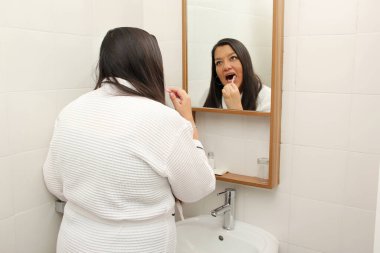 Latin 40 yaşında bir kadın banyoda aynanın önünde bornoz giymiş çürükleri önlemek için dişlerini fırçalıyor.