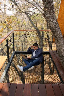 40 yaşındaki esmer tenli Latin bir adam evinin terasındaki ormanın üzerinde asılı duran bir ağ üzerinde dizüstü bilgisayarını kullanıyor ve tüm dünyada dijital göçebe olarak çalışıyor.