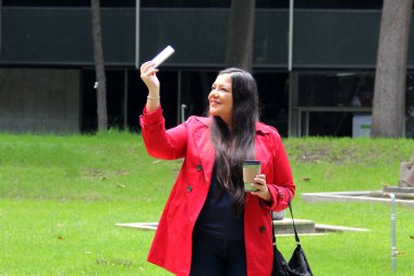 40 yaşında Latin bir kadın parkta kahve içerken ve cep telefonuyla çalışırken yürüyor, video görüşmeleri yapıyor, mesajlar gönderiyor ve ödemelerini yapıyor.