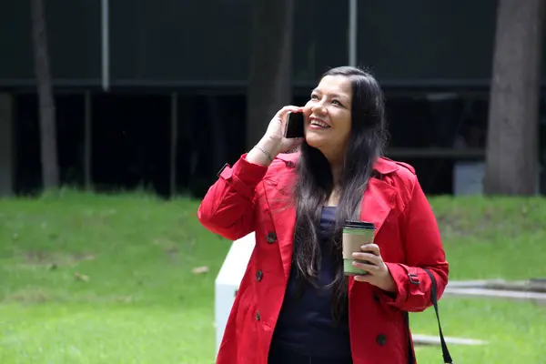 40-year-old Latina woman walks in the park while drinking coffee and using her cell phone to work, make video calls, send messages and make payments