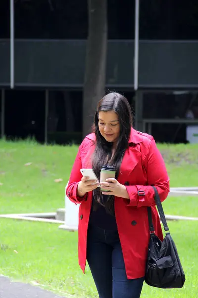 40-year-old Latina woman walks in the park while drinking coffee and using her cell phone to work, make video calls, send messages and make payments
