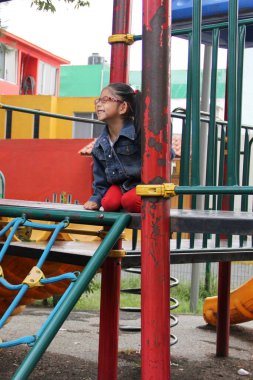 4 yaşındaki Latin esmer kız, gözlüklü, parkta oyun oynuyor. Yalnızlık ve yoksulluk çekiyor. Tatil günlerini otizmle geçiriyor.
