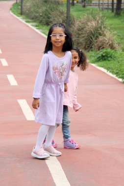 Brunette Latina 4 and 5 year old girls walk holding hands like sisters and friends spending time together outdoors clipart