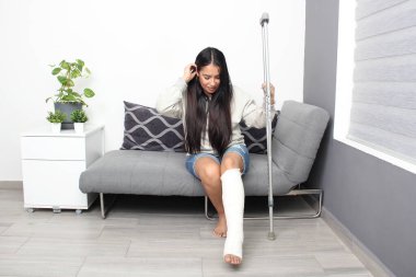 30 yaşında esmer bir kadın koltuk değneğiyle sandalyeden kalkmaya çalışıyor çünkü bacağı alçıda ve kazara kırılmış.