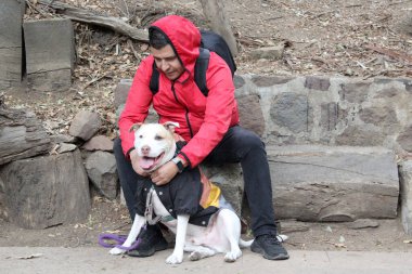 Koyu tenli Latin 40 yaşındaki adam yağmurlu bir günde parktaki bankta beyaz pitbull köpeğiyle oturuyor.