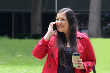 40 yaşında Latin bir kadın cep telefonuyla parkta yürüyor. Uzaktan çalışmak için, dışarıda video görüşmeleri yapıyor.