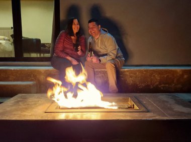 40 yaşındaki bir erkek ve kadın, aşklarını ve yıldönümlerini kutladıkları gece kamp ateşinin yanında sohbet ederken bira içerler.