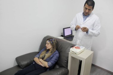 Esmer tenli Latin doktor, epilepsi teşhisi koymak için 30 yaşındaki bir kadının elektroensefalogram EEG 'si için telleri kafaya bağlıyor.