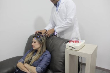 Esmer tenli Latin doktor, epilepsi teşhisi koymak için 30 yaşındaki bir kadının elektroensefalogram EEG 'si için telleri kafaya bağlıyor.