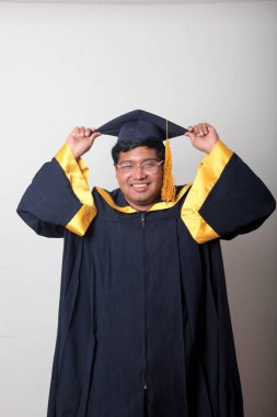 30 yaşında esmer tenli, gözlüklü bir adam mezuniyetini ve okulunu kutlamak için kep ve cüppe giyiyor.