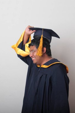 30 yaşında esmer tenli, gözlüklü bir adam mezuniyetini ve okulunu kutlamak için kep ve cüppe giyiyor.