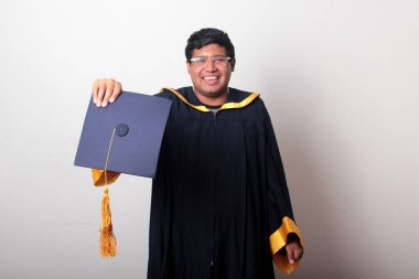 30 yaşında esmer tenli, gözlüklü bir adam mezuniyetini ve okulunu kutlamak için kep ve cüppe giyiyor.