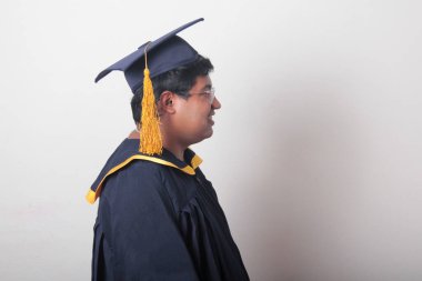 30 yaşında esmer tenli, gözlüklü bir adam mezuniyetini ve okulunu kutlamak için kep ve cüppe giyiyor.