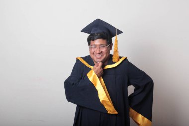 30 yaşında esmer tenli, gözlüklü bir adam mezuniyetini ve okulunu kutlamak için kep ve cüppe giyiyor.