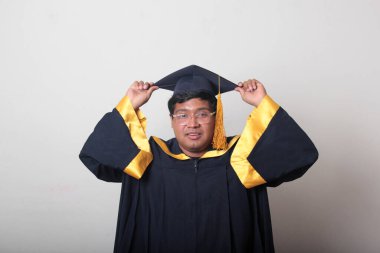 30 yaşında esmer tenli, gözlüklü bir adam mezuniyetini ve okulunu kutlamak için kep ve cüppe giyiyor.