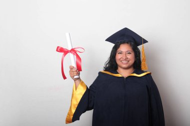 Şapka ve cübbe giymiş genç esmer kadın diplomasını gururla, tatmin olmuş ve kariyerini okulunda bitirmekten mutlu olarak gösteriyor.