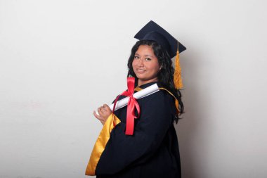 Şapka ve cübbe giymiş genç esmer kadın diplomasını gururla, tatmin olmuş ve kariyerini okulunda bitirmekten mutlu olarak gösteriyor.