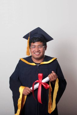 Başlık ve cübbe giyen genç adam gururla diplomasını gösteriyor. Mesleki eğitimini tamamlayıp mezun olmaktan memnun.