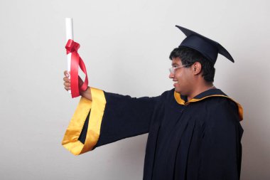 Başlık ve cübbe giyen genç adam gururla diplomasını gösteriyor. Mesleki eğitimini tamamlayıp mezun olmaktan memnun.