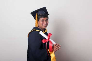 Başlık ve cübbe giyen genç adam gururla diplomasını gösteriyor. Mesleki eğitimini tamamlayıp mezun olmaktan memnun.