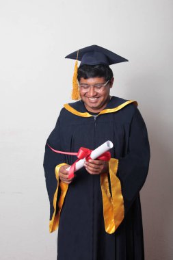 Başlık ve cübbe giyen genç adam gururla diplomasını gösteriyor. Mesleki eğitimini tamamlayıp mezun olmaktan memnun.