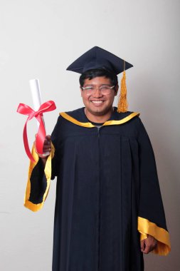 Başlık ve cübbe giyen genç adam gururla diplomasını gösteriyor. Mesleki eğitimini tamamlayıp mezun olmaktan memnun.