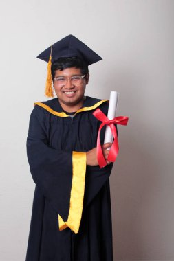 Başlık ve cübbe giyen genç adam gururla diplomasını gösteriyor. Mesleki eğitimini tamamlayıp mezun olmaktan memnun.