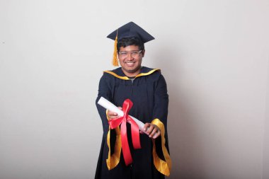 Başlık ve cübbe giyen genç adam gururla diplomasını gösteriyor. Mesleki eğitimini tamamlayıp mezun olmaktan memnun.