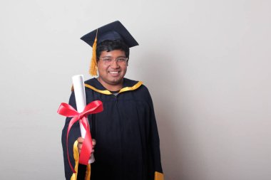 Başlık ve cübbe giyen genç adam gururla diplomasını gösteriyor. Mesleki eğitimini tamamlayıp mezun olmaktan memnun.