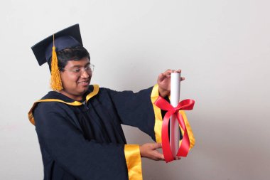 Başlık ve cübbe giyen genç adam gururla diplomasını gösteriyor. Mesleki eğitimini tamamlayıp mezun olmaktan memnun.