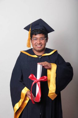 Başlık ve cübbe giyen genç adam gururla diplomasını gösteriyor. Mesleki eğitimini tamamlayıp mezun olmaktan memnun.