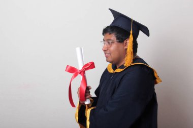 Başlık ve cübbe giyen genç adam gururla diplomasını gösteriyor. Mesleki eğitimini tamamlayıp mezun olmaktan memnun.