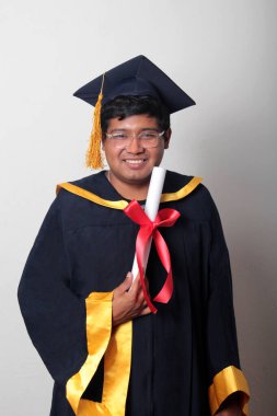 Başlık ve cübbe giyen genç adam gururla diplomasını gösteriyor. Mesleki eğitimini tamamlayıp mezun olmaktan memnun.