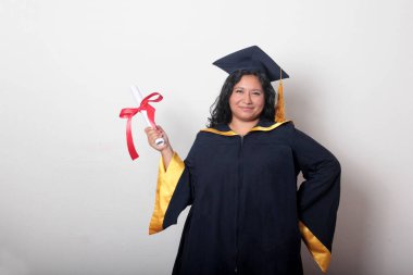 Şapka ve cübbe giymiş genç esmer kadın diplomasını gururla, tatmin olmuş ve kariyerini okulunda bitirmekten mutlu olarak gösteriyor.