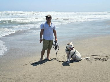 Yetişkin bir adam, plajda terapi olarak beyaz pitbull 'uyla yürüyor ve yanında depresyon ve anksiyeteye karşı beslediği duygusal destek de var.