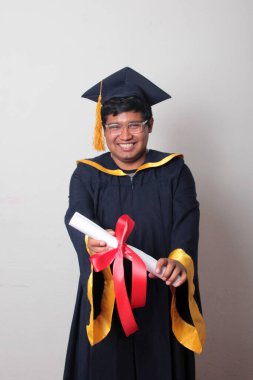 Başlık ve cübbe giyen genç adam gururla diplomasını gösteriyor. Mesleki eğitimini tamamlayıp mezun olmaktan memnun.