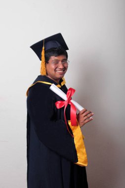 Başlık ve cübbe giyen genç adam gururla diplomasını gösteriyor. Mesleki eğitimini tamamlayıp mezun olmaktan memnun.