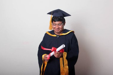 Başlık ve cübbe giyen genç adam gururla diplomasını gösteriyor. Mesleki eğitimini tamamlayıp mezun olmaktan memnun.