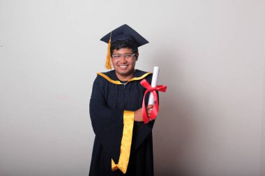 Başlık ve cübbe giyen genç adam gururla diplomasını gösteriyor. Mesleki eğitimini tamamlayıp mezun olmaktan memnun.