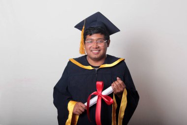 Başlık ve cübbe giyen genç adam gururla diplomasını gösteriyor. Mesleki eğitimini tamamlayıp mezun olmaktan memnun.