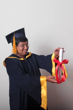 Başlık ve cübbe giyen genç adam gururla diplomasını gösteriyor. Mesleki eğitimini tamamlayıp mezun olmaktan memnun.