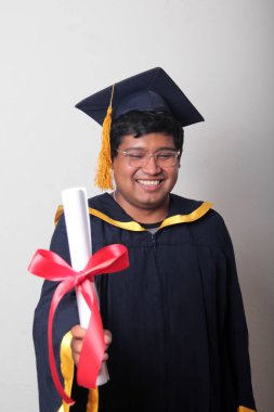 Başlık ve cübbe giyen genç adam gururla diplomasını gösteriyor. Mesleki eğitimini tamamlayıp mezun olmaktan memnun.