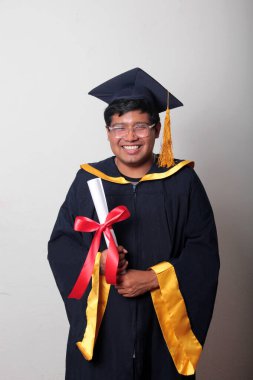 Başlık ve cübbe giyen genç adam gururla diplomasını gösteriyor. Mesleki eğitimini tamamlayıp mezun olmaktan memnun.