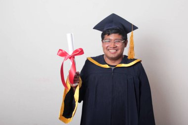 Başlık ve cübbe giyen genç adam gururla diplomasını gösteriyor. Mesleki eğitimini tamamlayıp mezun olmaktan memnun.