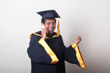 Mezuniyetini kutlamak için kep ve cübbe giymiş genç bir adam cep telefonunu selfie çekmek, arama yapmak ve baloda mesaj göndermek için kullanıyor.