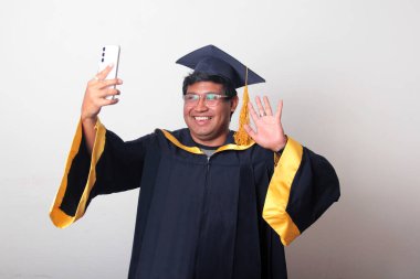 Mezuniyetini kutlamak için kep ve cübbe giymiş genç bir adam cep telefonunu selfie çekmek, arama yapmak ve baloda mesaj göndermek için kullanıyor.