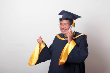 Mezuniyetini kutlamak için kep ve cübbe giymiş genç bir adam cep telefonunu selfie çekmek, arama yapmak ve baloda mesaj göndermek için kullanıyor.